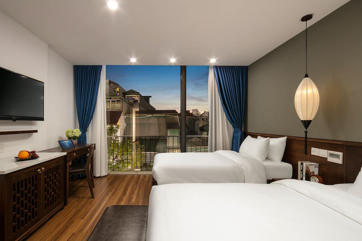 A hotel room with two beds, a desk with a laptop, a TV, and a balcony with curtains.