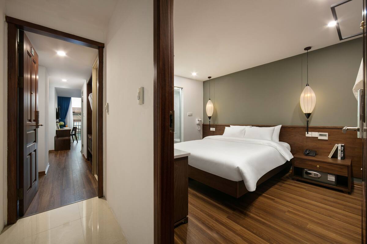 A hotel room with a wooden floor, a large bed with white linens, a bedside table with a lamp, and a view of the hallway.
