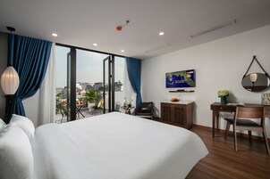 A modern hotel room with a large bed, a desk, and a chair. There is a television on the wall and a view of the city through the window.