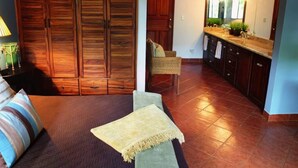 A room with a bed, a chair, a wooden wardrobe, a kitchen area with a sink and mirror, and a tiled floor.