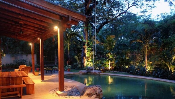 A pool area with a wooden pergola, seating, and tropical vegetation.