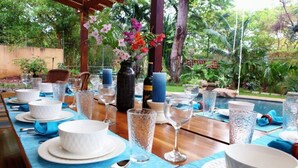 A dining table set for a meal with white plates, bowls, and clear glasses, surrounded by a lush garden view.
