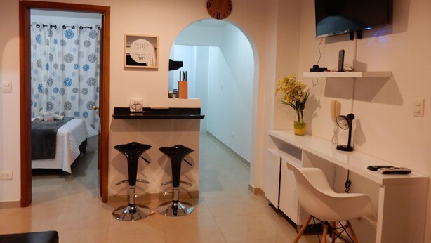 A modern kitchen with a central island, two bar stools, a dining table, and a TV mounted on the wall.