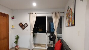A living room with a black sofa, red cushions, a red rug, a potted plant, a standing fan, and a window with curtains.