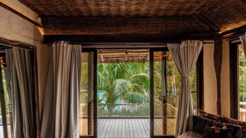 Una habitación con techo de madera, una puerta de cristal corredera que da a un balcón con piscina y palmeras, y una zona de estar con cojines.
