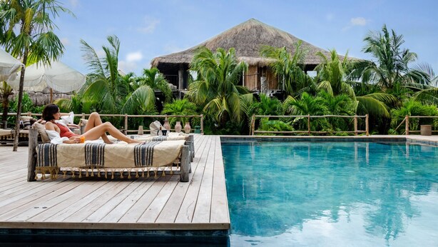 A poolside area with a wooden deck, lounge chairs, and a thatched-roof building in the background. - Radhoo Tulum (Tulum)