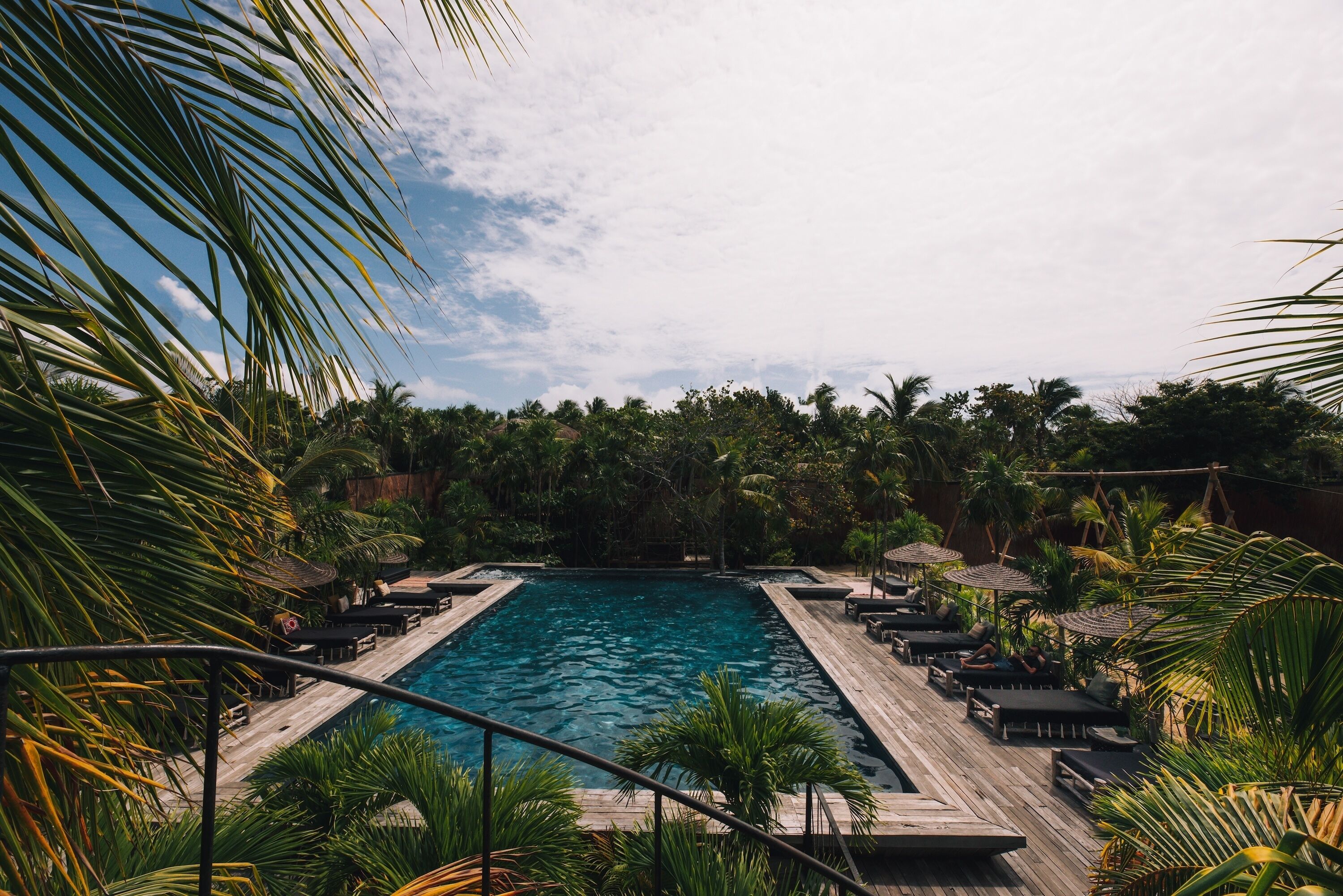 Piscina retangular com deck de madeira e espreguiçadeiras, cercada por vegetação exuberante e palmeiras.