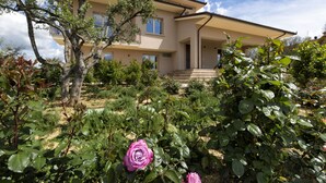 Une maison à deux étages, à la façade beige, dotée d’un porche et d’un jardin où poussent différentes plantes, ainsi qu’un rosier rose.