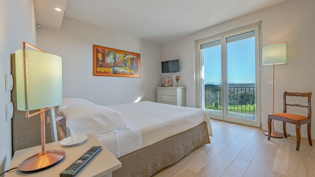 Une chambre d’hôtel avec un lit, un bureau doté d’une lampe, une télécommande, un téléviseur posé sur un meuble, une chaise et un balcon avec vue.