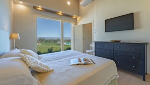 A bedroom with a large bed, a dresser, a flat-screen TV, and a view of a green landscape through a large window.
