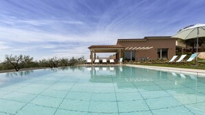 A swimming pool with lounge chairs and a covered seating area, surrounded by a landscaped garden and a building with large windows.