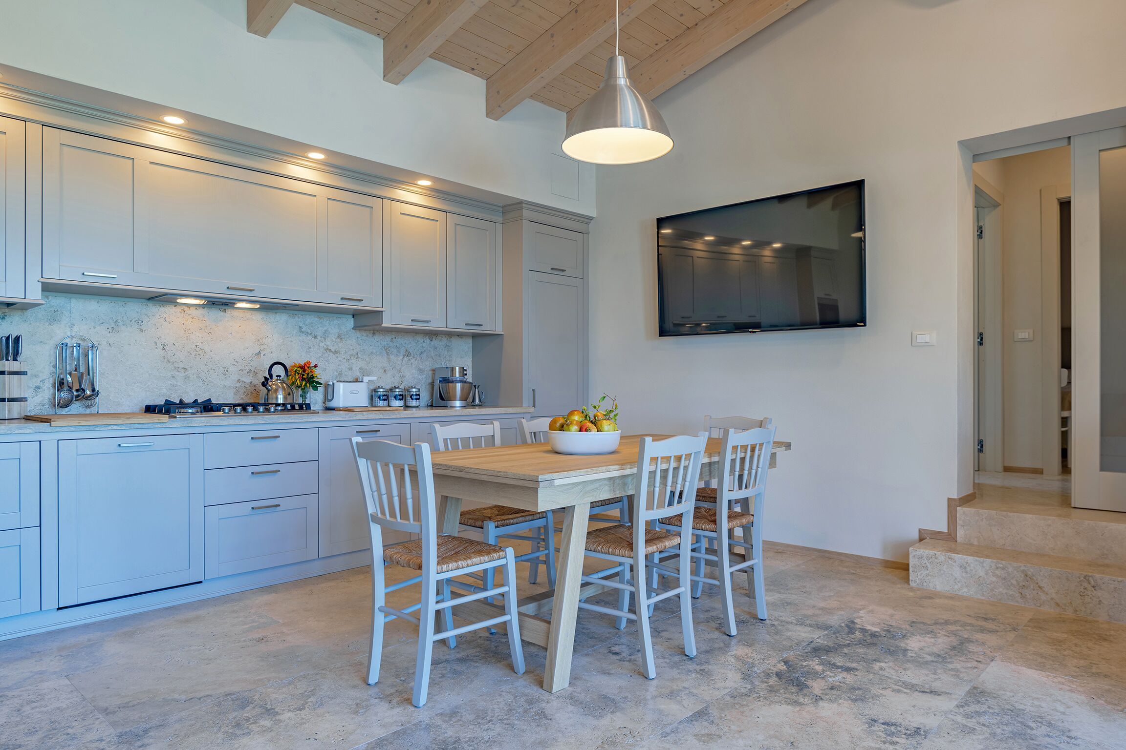 A modern kitchen with white cabinets, a wooden dining table, and a large TV mounted on the wall.
