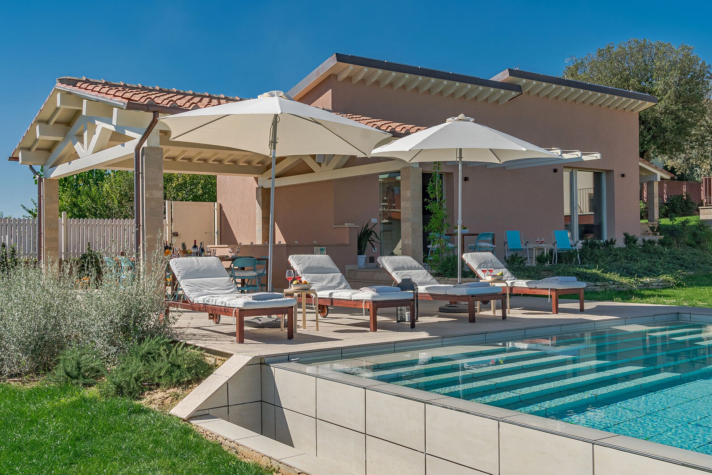 Un espace aménagé au bord de la piscine, avec des chaises longues et des parasols, une piscine et un bâtiment doté d’un toit recouvert de tuiles.
