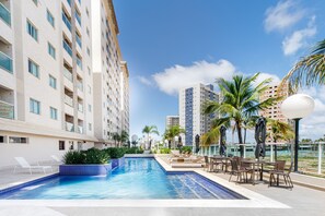 Una piscina con sillas de descanso y mesas, rodeada de edificios altos y palmeras.