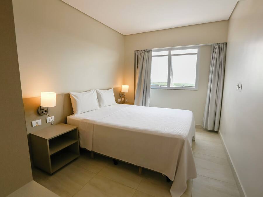 A hotel room with a bed, nightstands, a lamp, a window with curtains, and a view of greenery outside.
