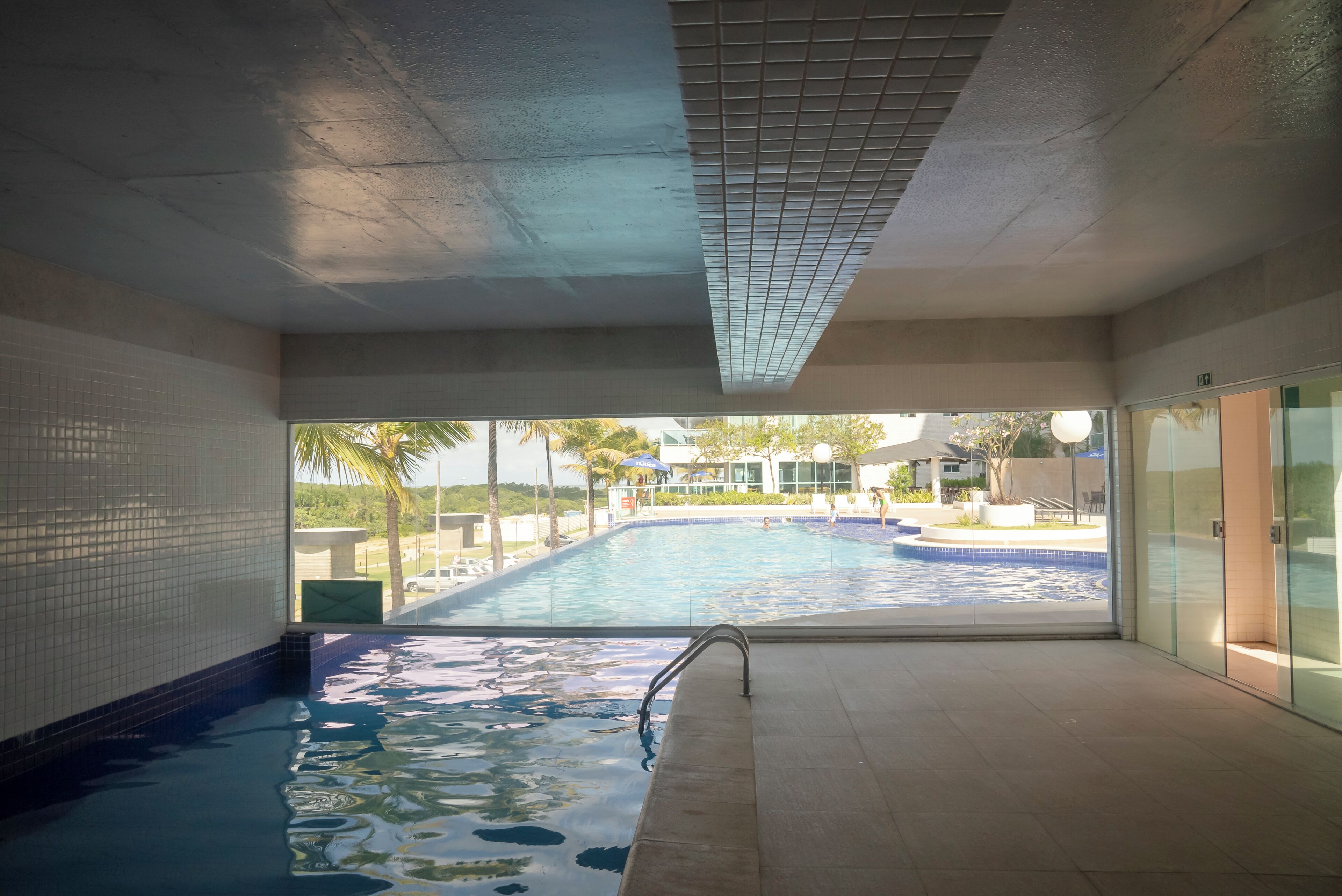 An indoor swimming pool with clear glass walls, a tiled floor, and a view of an outdoor pool area with palm trees.