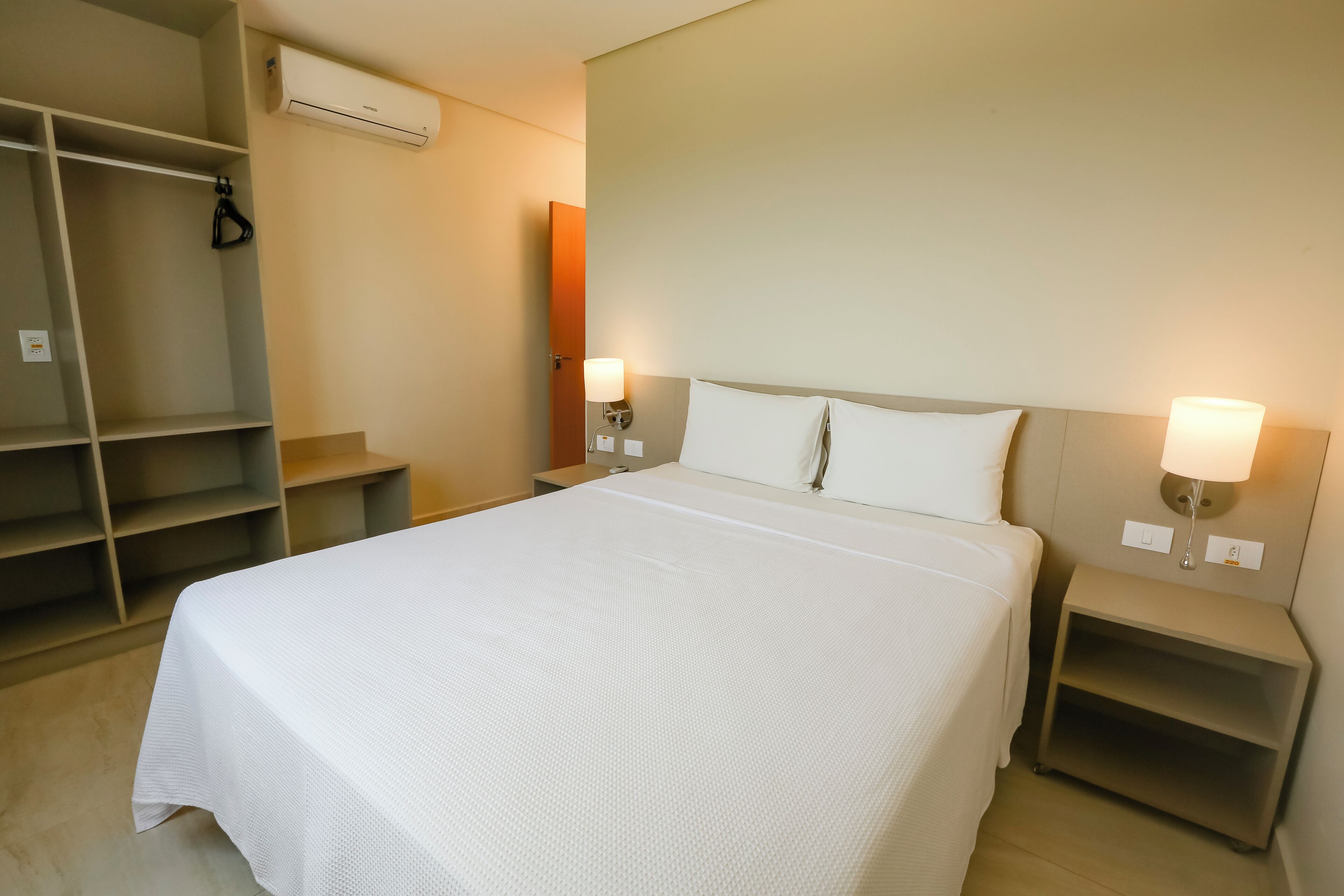 A neatly arranged hotel room with a large bed, two bedside tables with lamps, a wardrobe, and an air conditioning unit.