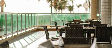 A covered outdoor seating area with wicker furniture and a glass balustrade overlooking a garden with palm trees.
