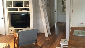 Una habitación con piso de madera, televisión, silla, escalera y mesa con un letrero.