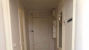 Un pasillo estrecho con piso de madera que conduce a una puerta blanca. A la izquierda, hay una pequeña mesa redonda con una planta y un cesto de basura.