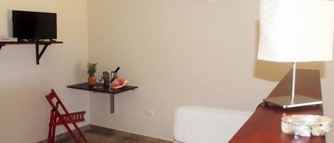 Une petite pièce avec un téléviseur posé sur une étagère, un fauteuil rouge, une petite table avec une lampe et un climatiseur fixé au mur.