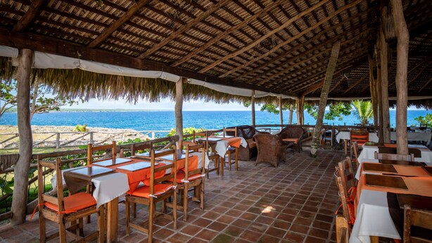 An outdoor dining area with wooden tables and chairs, a thatched roof, and a view of the ocean. - Hotel Bocaino (San Rafael del Yuma)