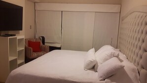 A hotel room with a large bed, a television on a stand, a striped armchair, and a wardrobe with curtains. - GL Apts, rent Upper Pardo Miraflores (Miraflores)