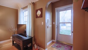 A room with a wooden floor, a dark wooden desk with drawers, a flat-screen TV, a vase with branches, and a door with a glass panel. - Charming home near Boise State and The Train Depot
 (Boise)