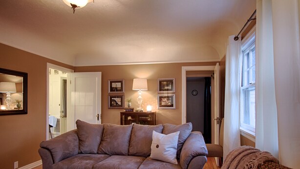 A living room with a brown sofa, a coffee table with magazines, and a lamp. There is a door leading to another room and a window with curtains. - Charming home near Boise State and The Train Depot
 (Boise)