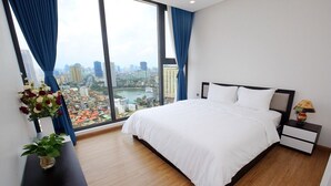 A modern hotel room with a large bed, a nightstand, a vase with flowers, and a view of a cityscape through large windows. - 3 bedroom in Metropolis near Lotte, Dao Tan (Hanoi)