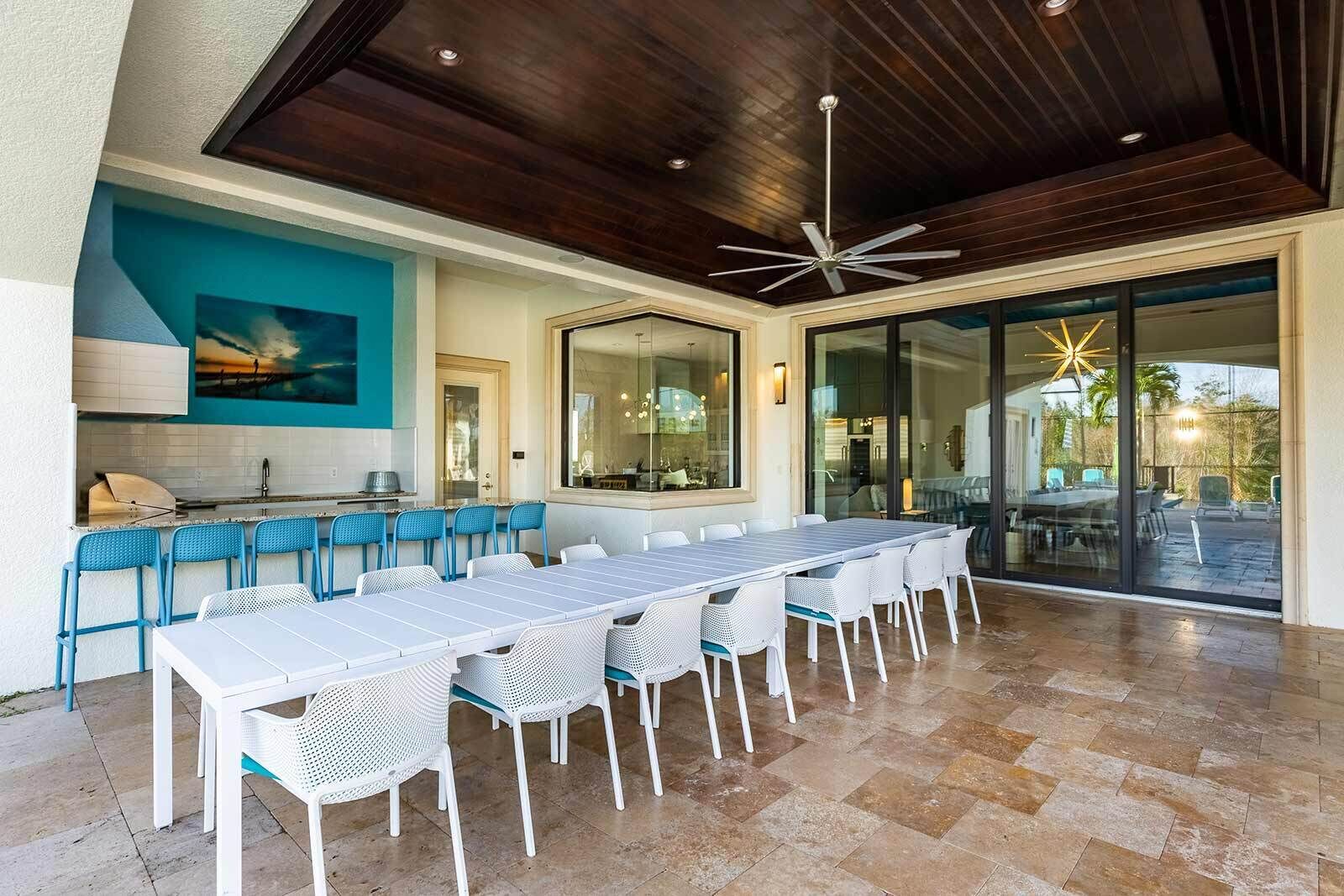 An outdoor dining area with a long table and chairs, a kitchen area with a blue backsplash, and a ceiling fan.