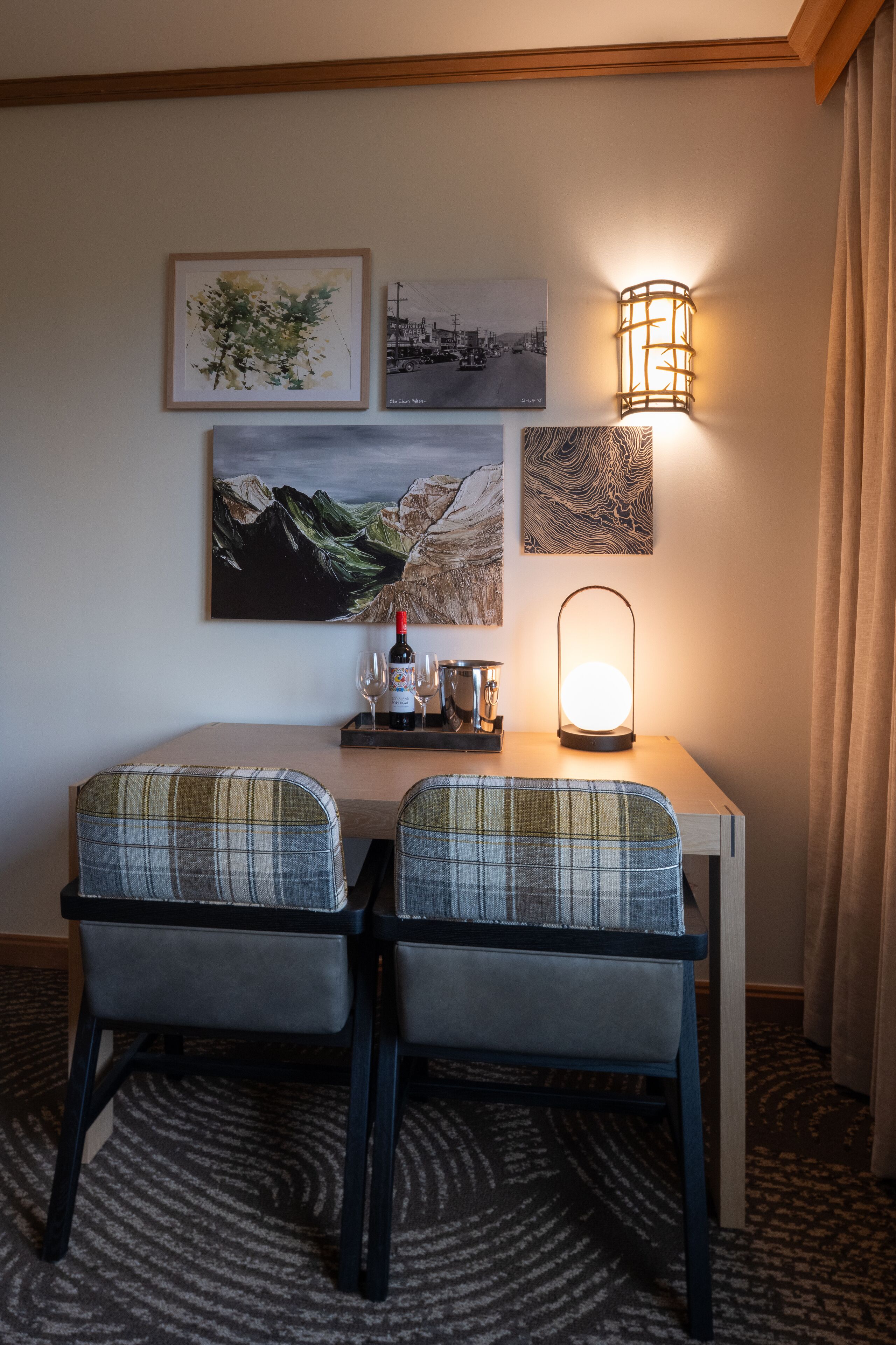 Un rincón de comedor con dos sillones a cuadros, una mesa con una botella de vino, dos copas de vino y una lámpara de pared.
