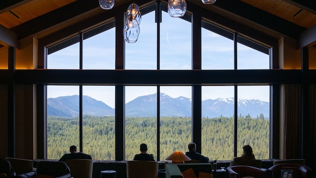 Una habitación amplia con ventanas grandes que ofrecen vistas a la montaña, vigas de madera en el techo y lámparas colgantes.