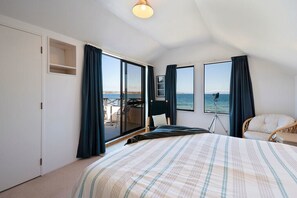 A bedroom with a large bed, a desk, and a chair, with a view of the sea through the windows.