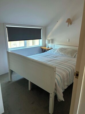 Une chambre à coucher avec un lit, une fenêtre avec un store, une table de chevet avec une lampe et un luminaire fixé au mur.