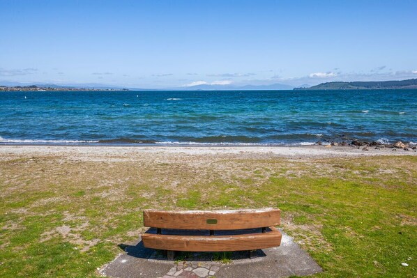 A wooden bench on a concrete platform by a beach with a view of the sea and distant hills.