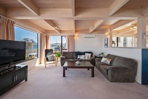 Ein Wohnzimmer mit großem Flachbildfernseher, einer Couch, einem Couchtisch und Blick ins Freie.