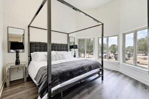A four-poster bed with a dark headboard, a bedside table with a lamp, and large windows offering a view of the outside. - 9000 Sqft Estate/Pickleball/Basketball/Pool/HotTub/9 Bdrm/8 Bath/23Beds/Sleeps35 (Dripping Springs)