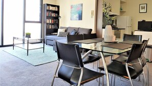 A modern living room with a glass dining table, black chairs, a grey sofa, a bookshelf, and a kitchen area in the background. - In the heart of Southbank! (Southbank)