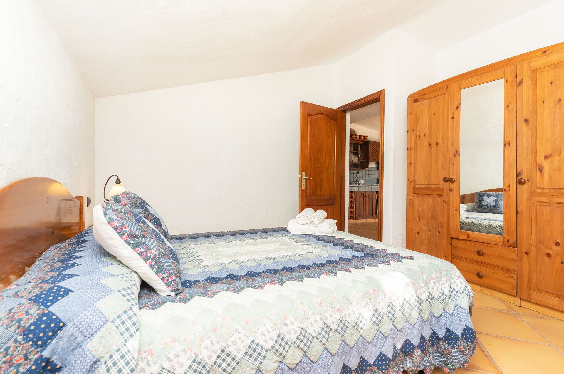 A bedroom with a bed, a wooden wardrobe, and a door leading to a kitchen.