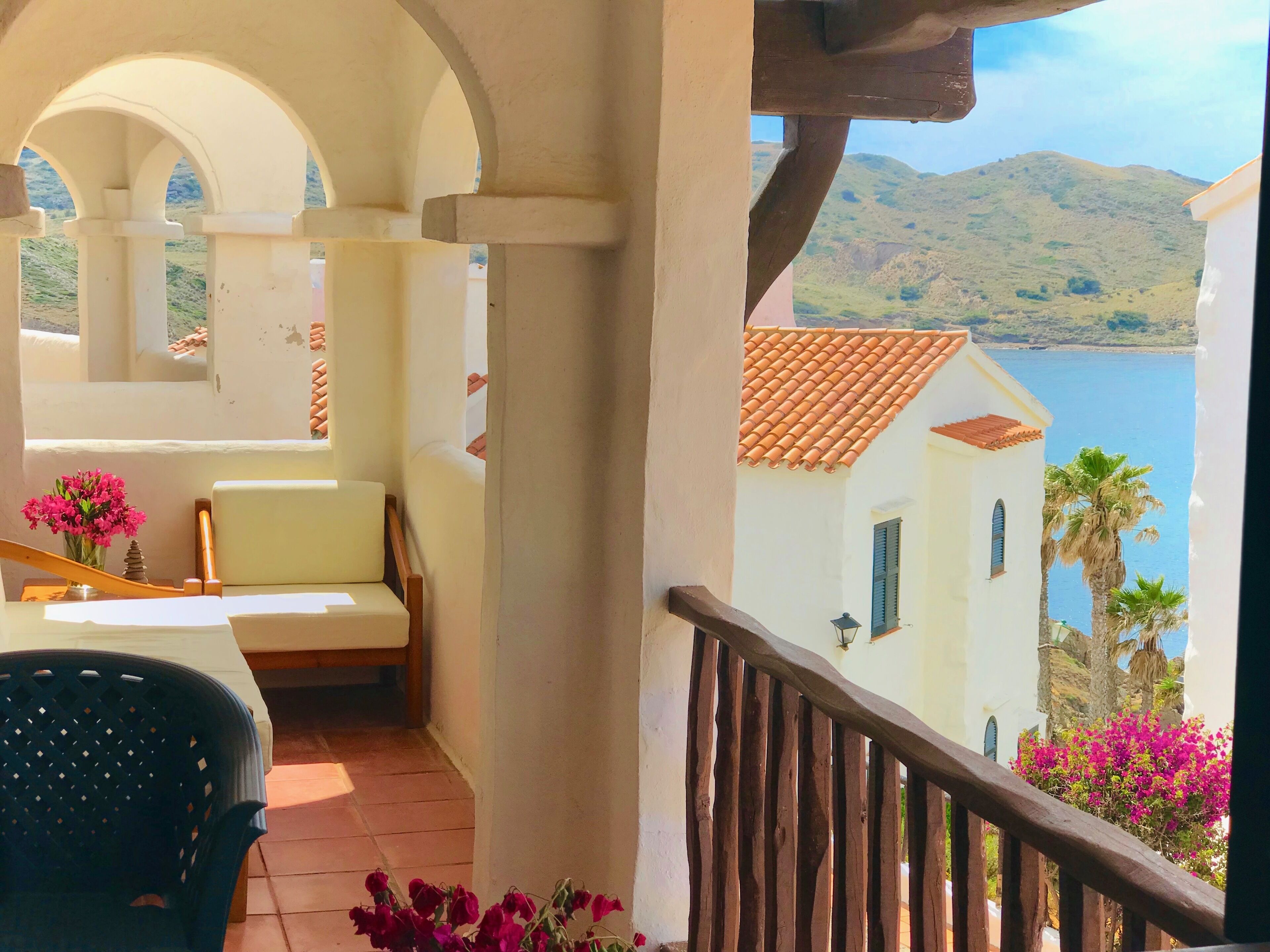 A balcony with a white chair and a table, overlooking a body of water and hills.
