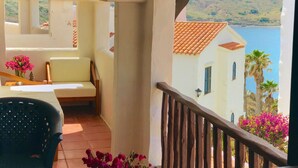 Un balcon avec une chaise blanche et une table, donnant sur une étendue d’eau et des collines.