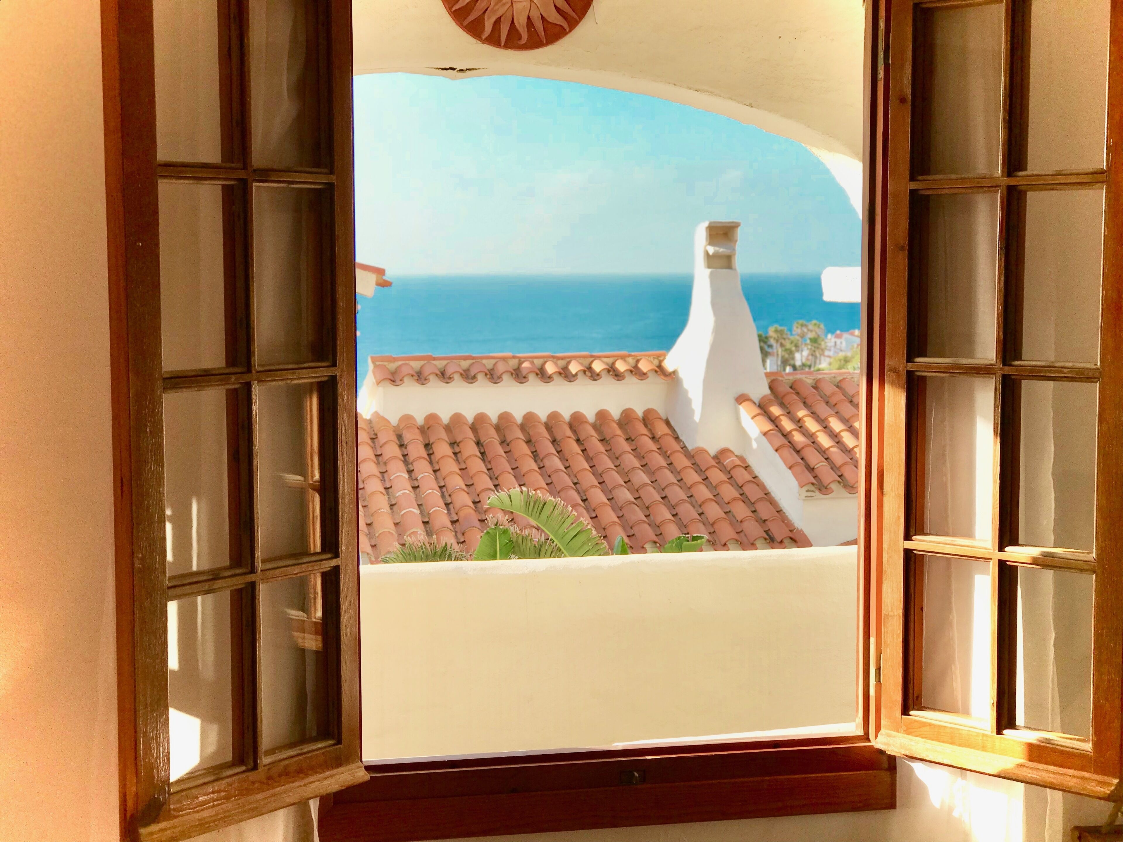 A view through a window with a tiled roof and a white wall, overlooking a coastal area with a clear sky and the sea.