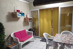 A small, enclosed patio area with a white plastic chair, a table with a patterned tablecloth, a small shelf with towels and toiletries, and a sliding glass door with a yellow curtain.