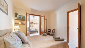 A bedroom with a bed, a bedside table with a lamp, a chair, a wooden door, and a shelf with decorative items. - Apartment Carla near the beach with spacious balcony, mountain view, Wi-Fi & air conditioning (Puerto de Soller)