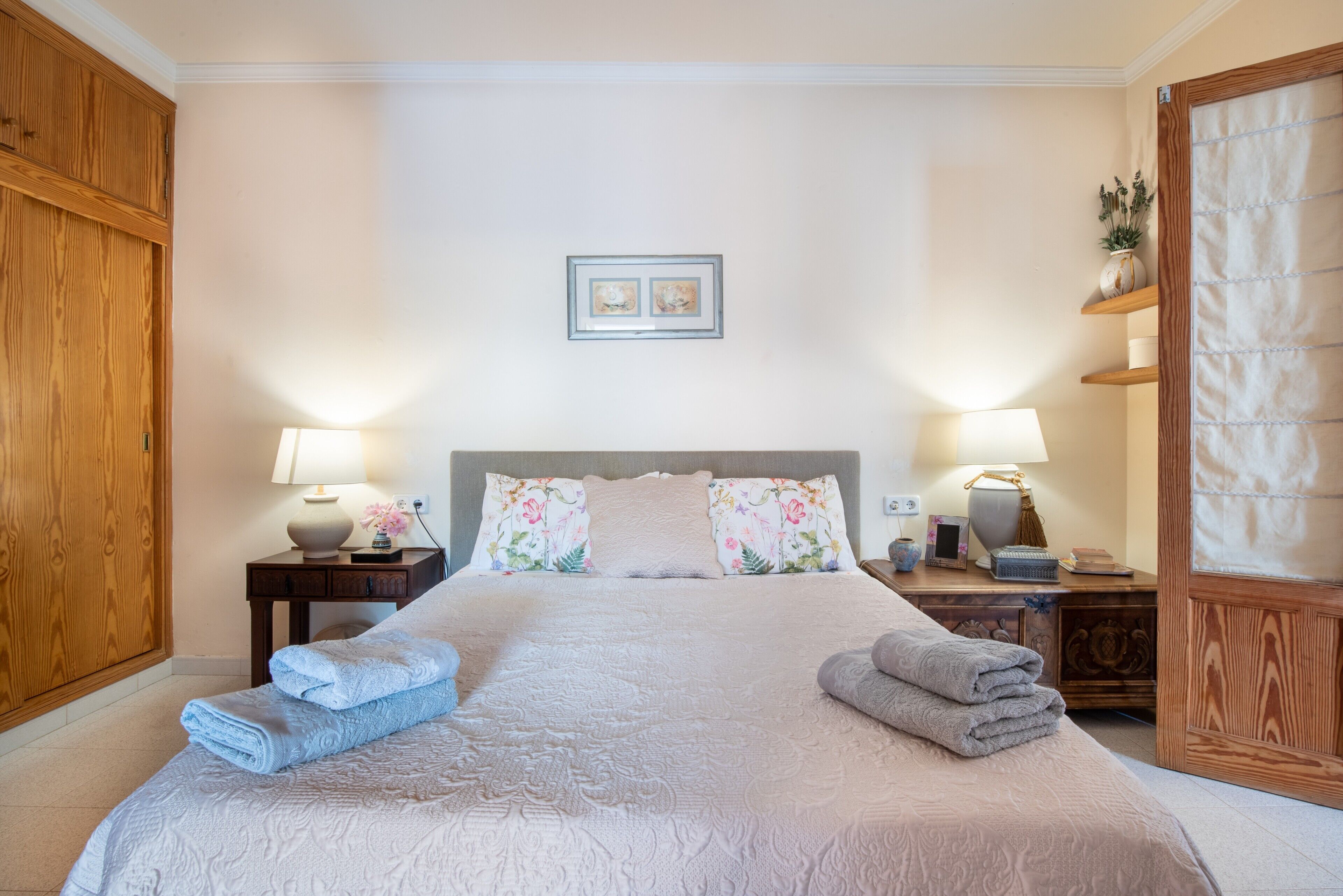Una cama bien hecha con almohadas y toallas dobladas, una mesita de noche con lámpara y un armario de madera.