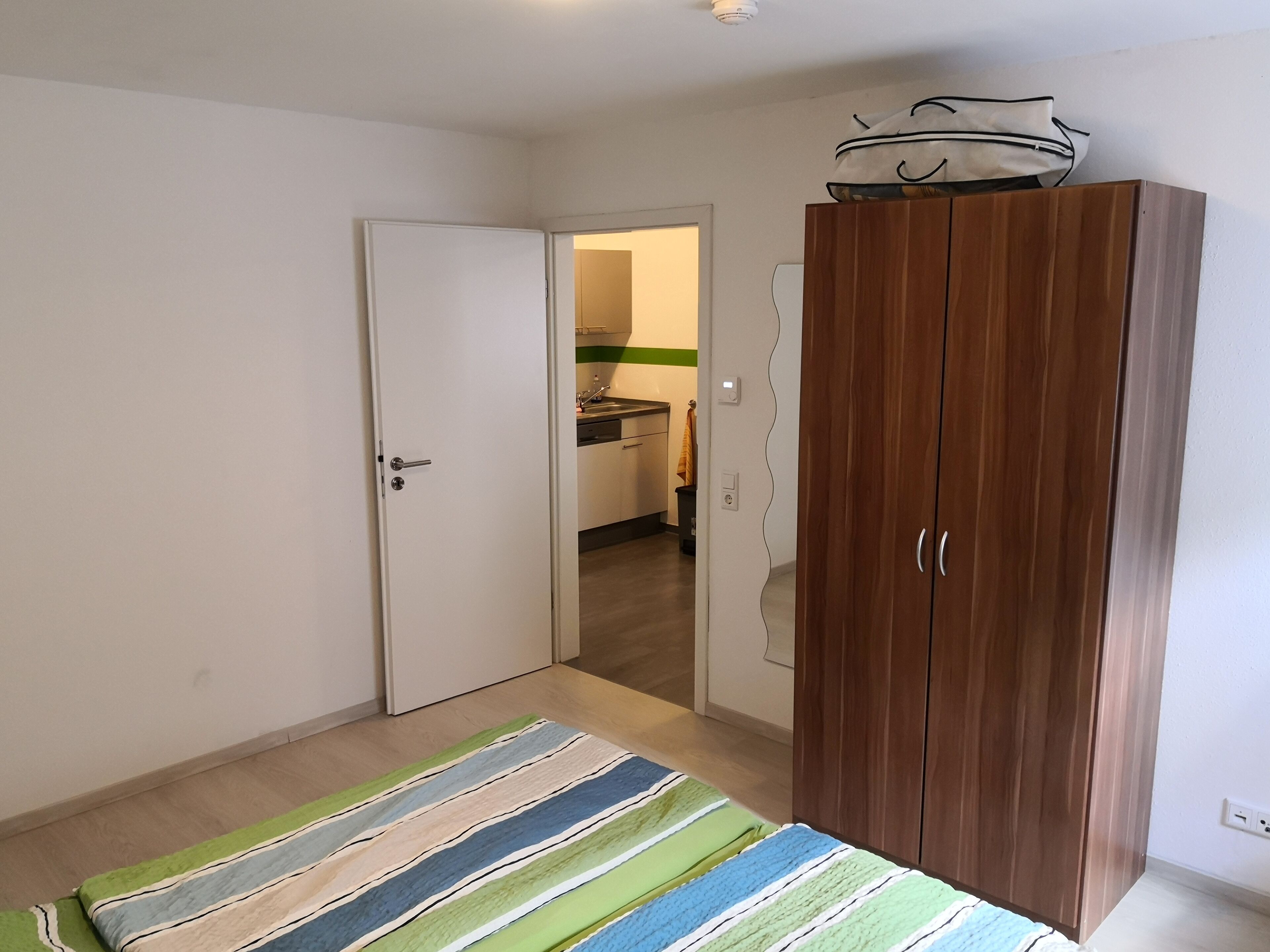 A room with a wooden wardrobe, a striped rug, and a door leading to a kitchen.