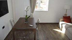 Un salon moderne avec un coin repas, comprenant une table en bois, des chaises transparentes, une orchidée en pot, un téléviseur fixé au mur et une vue sur la ville par une fenêtre.