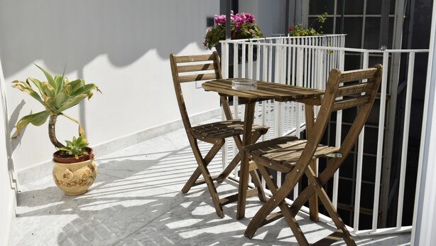 Une table et des chaises en bois sur un balcon doté d’une balustrade blanche, une plante en pot et une fenêtre.