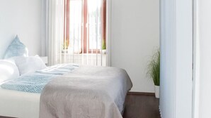 Een slaapkamer met een bed, witte muren, een raam met zonwering en een potplant.